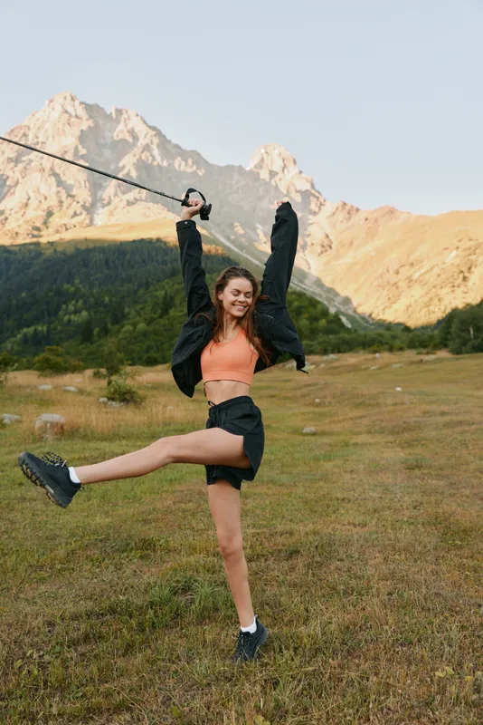 A young woman enjoys an energetic outdoor moment, standing in a grassy valley with rugged mountains in the background. She wears sporty clothes, raises arms in triumph, and smiles brightly.