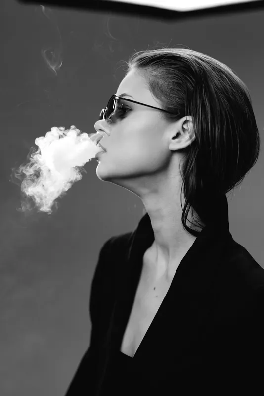 Black and white portrait of a woman smoking, wearing sunglasses, exhaling smoke with a calm expression.
