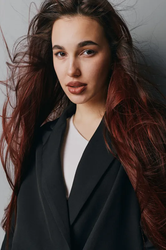 Woman with long red hair and natural makeup wearing black coat and white top posing against gray background in studio. Portrait of confident young female with loose hairstyle.