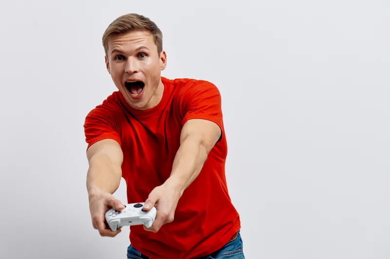 Energetic young man with short blond hair wearing bright red t-shirt plays video game with game controller surprised excited expression on a plain white background. People lifestyle concept