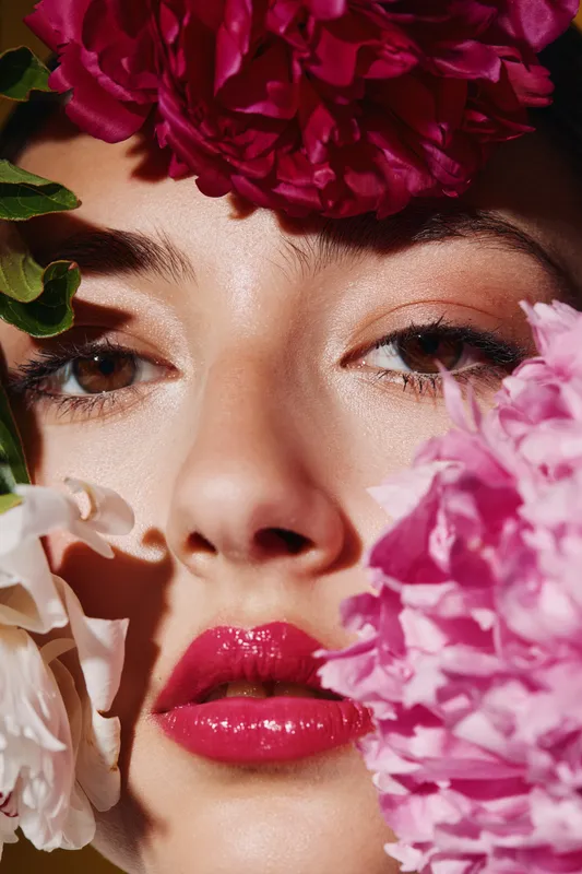 Close-up portrait of woman with glossy lips surrounded by vibrant pink and red flowers. Creative beauty fashion shot with soft natural lighting and fresh spring vibes.