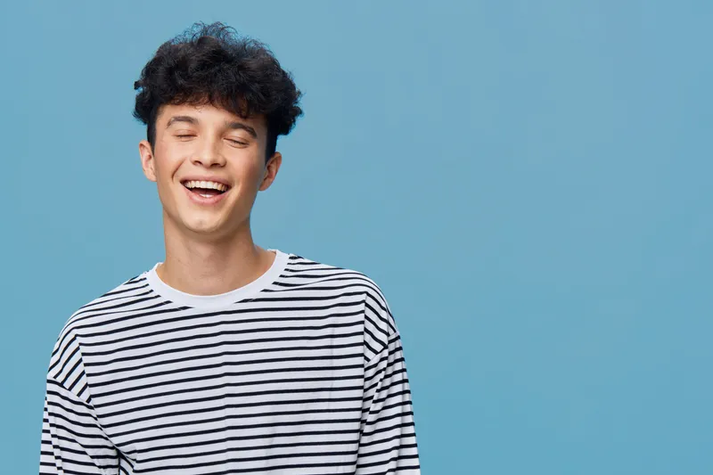 smiling man in striped shirt enjoying joyful moment with closed eyes, curly hair, and blue background expressing happiness and relaxation in casual style