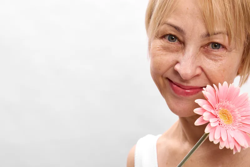 Smiling woman holding a pink flower on a stick, closeup portrait on white background, skincare and wellness concept, soft studio lighting, gentle happy mood
