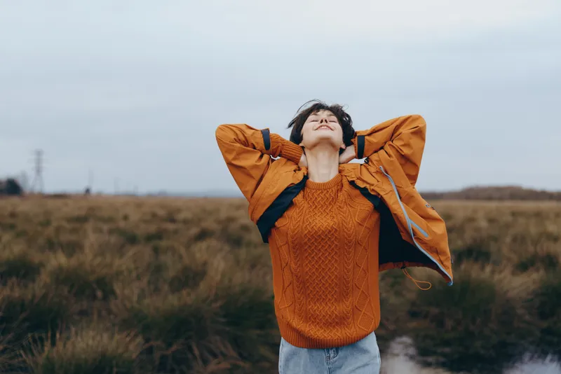 Woman lifestyle outdoors, laughing with eyes closed, orange sweater and casual jacket, standing on a grassy path, wind moving hair, cloudy sky, calm mood, natural light, wide field open air