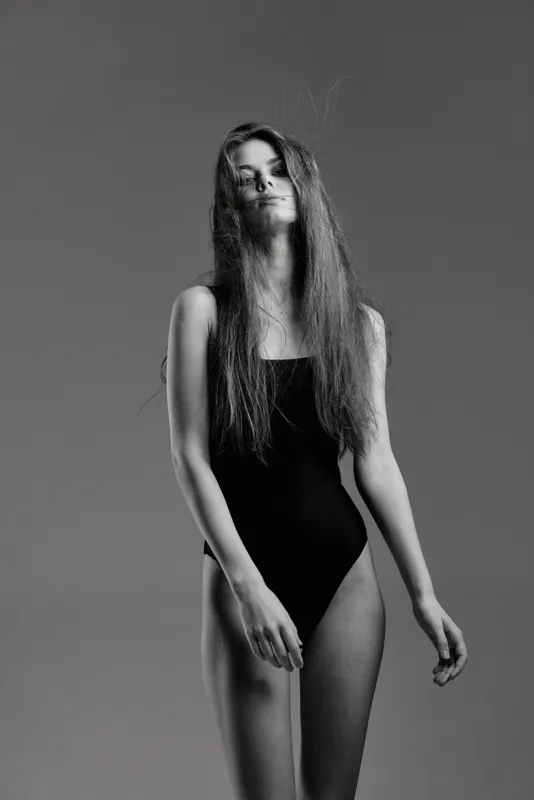 Black and white portrait of a tall woman with long hair wearing a black bodysuit posing against gray background.