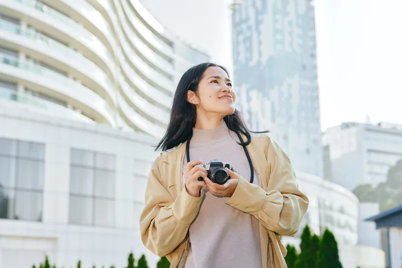 Camera, woman, outdoors, urban, city scene with daylight, a casual jacketed figure holds a camera and gazes skyward amid modern buildings and a bright skyline, capturing a candid travel moment
