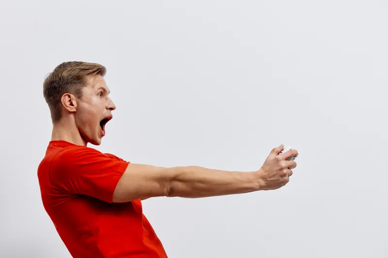 Excited young man with short brown hair and light skin wearing a bright red t-shirt shouting and expressing surprise with an open mouth and wide eyes, holding a smartphone in his outstretched hand aga