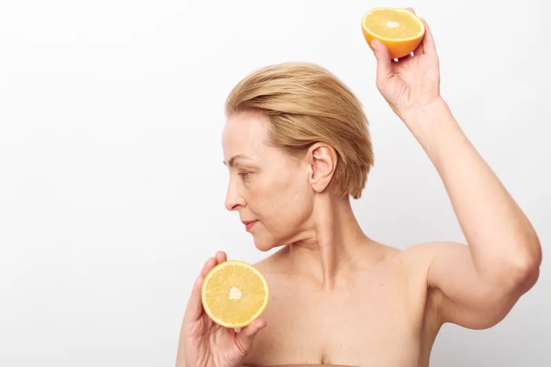 Mature woman with short blonde hair holds two halves of citrus fruit near her face and shoulder, isolated on white background. Concept of natural skincare, wellness, and healthy lifestyle.