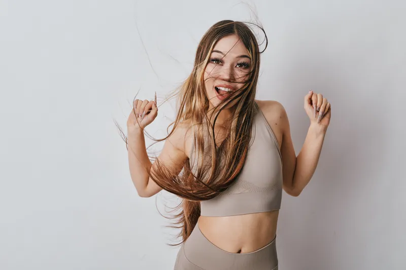 Young woman with long hair dancing and smiling in casual sportswear for fitness, joy and movement on plain background. Female model with energetic pose in studio for lifestyle and health.