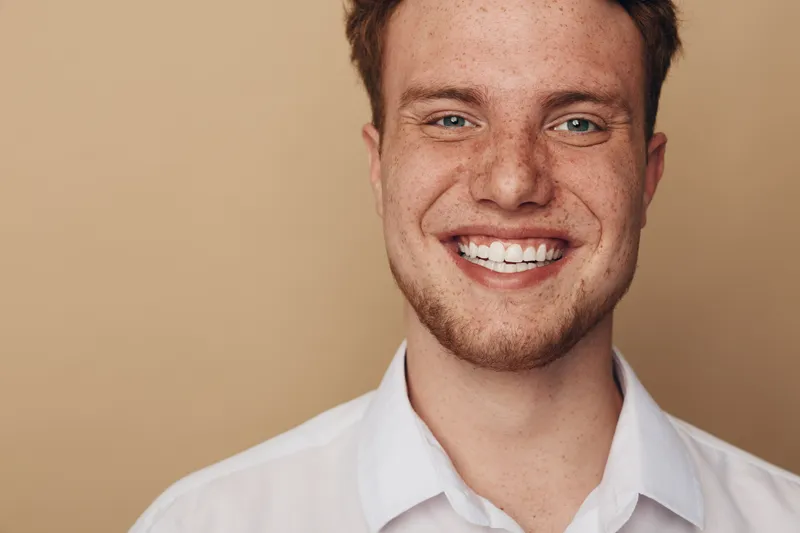 Smiling young man with freckles and red hair wears white shirt against beige background. Close-up portrait with natural light and happy positive expression. Concept of youth and confidence.