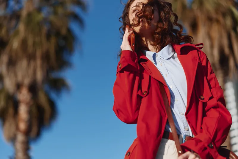 A young woman in a red coat and light blue shirt stands outdoors under a bright blue sky, looking off to the side with a thoughtful expression.