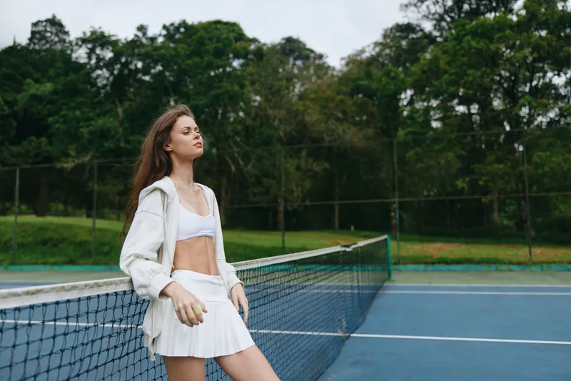 Young woman in sporty attire enjoying a moment of tranquility on a tennis court surrounded by greenery, embodying athleticism and confidence