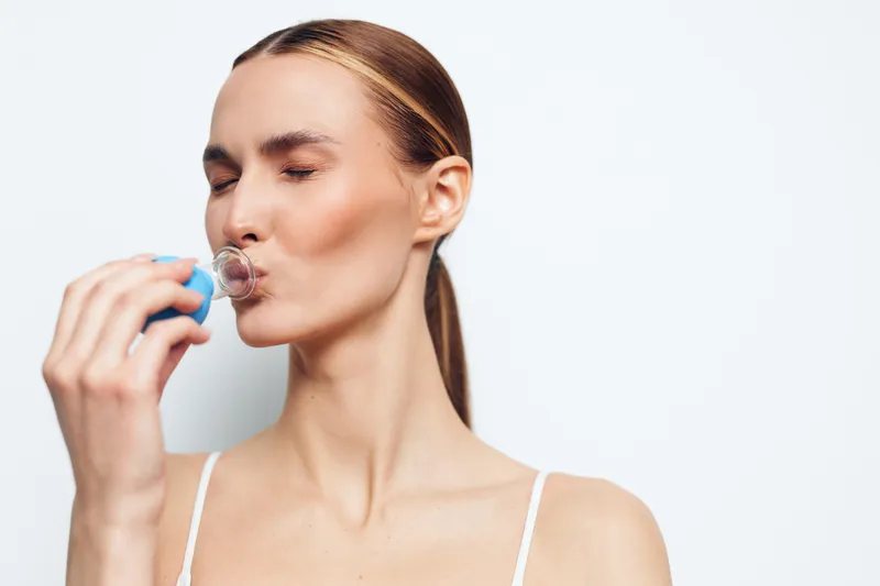 Young woman with a ponytail wearing a white tank top holds a blue spray bottle near her lips, performing a close up skincare moment in a clean studio setting.