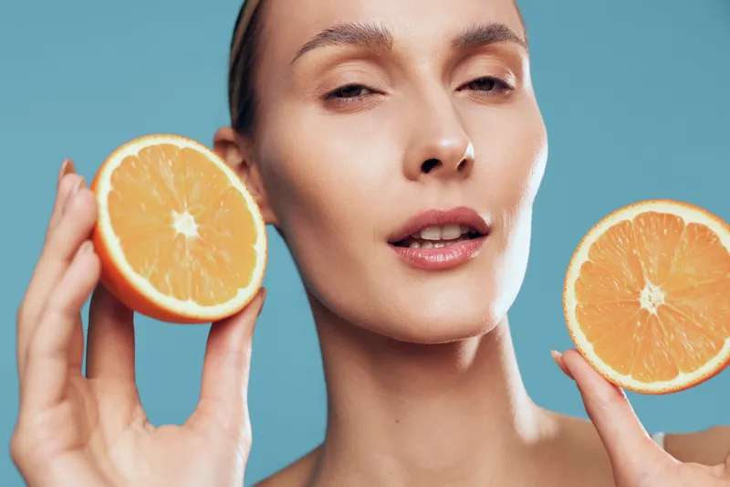 Young woman holds two halves of fresh orange fruit near face with calm expression isolated on blue background, healthy lifestyle concept and natural beauty close up portrait.