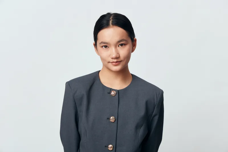 Young woman with black hair and elegant gray blazer posing with neutral expression on white background for portrait, confidence, and professional appearance in studio lighting.