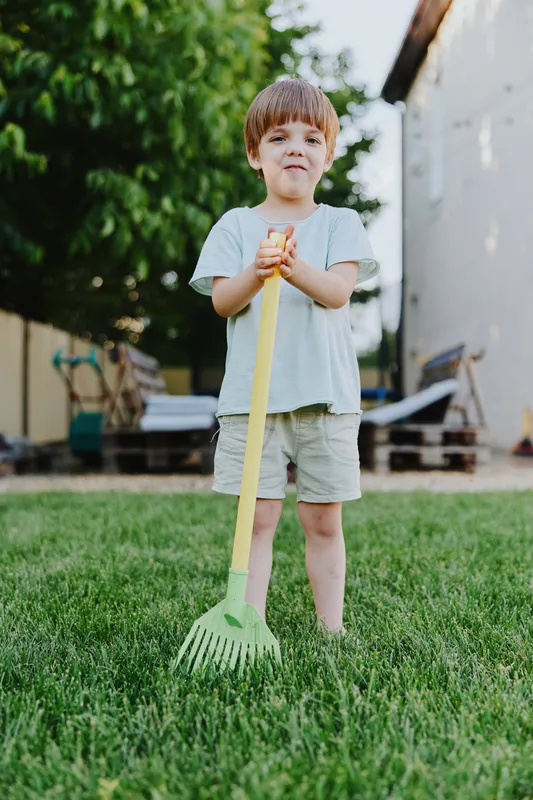 Young boy holding a colorful rake, standing on lush green grass, smiling with joy. This playful moment captures the essence of childhood and outdoor fun.