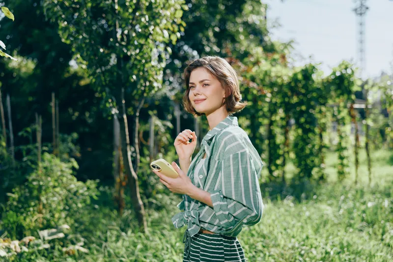 Woman, outdoor, nature, smile a cheerful woman stands in a sunny garden with a phone in her hand, radiating confidence, happiness, and readiness for a peaceful day outdoors.