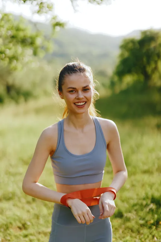 Young athletic woman in gray sports wear smiling while exercising outdoors with resistance bands in a green natural setting during a sunny day