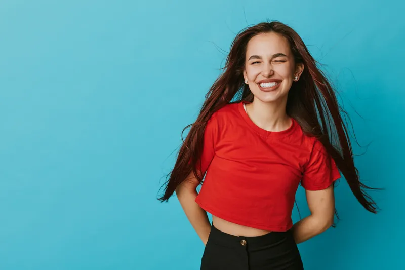 Young woman smiling with eyes closed wearing red shirt and black pants standing against blue background. Happy female with long hair and casual outfit in studio portrait with natural expression.