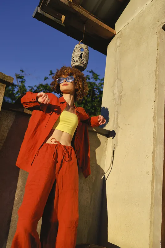 A woman in bold red-orange outfit and yellow top poses outdoors, leaning against a sunlit wall with a hanging lantern above, a lifestyle portrait of confidence and fashion