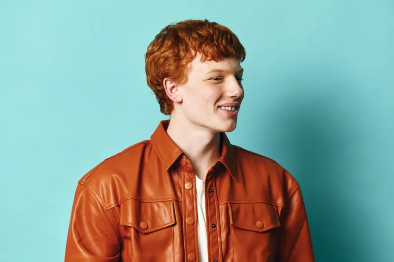 Young man with red hair wearing a brown leather jacket smiling while looking away on blue background for casual style and fashion portrait photo session.