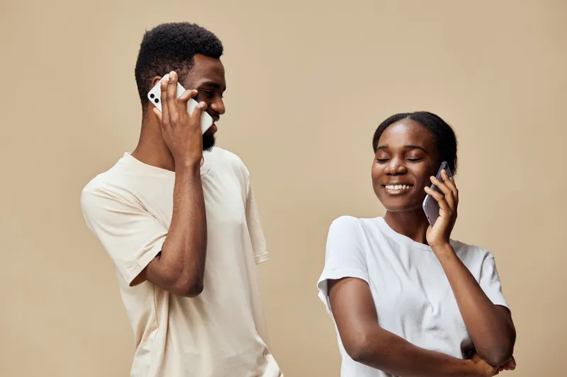 Happy young African couple enjoying a fun phone conversation against a neutral beige background, dressed casually in matching light colored clothing