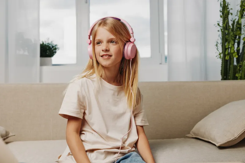 Blond girl with pink headphones sits on beige sofa in bright room, looking away and smiling. Concept for childhood, music, and relaxation at home.