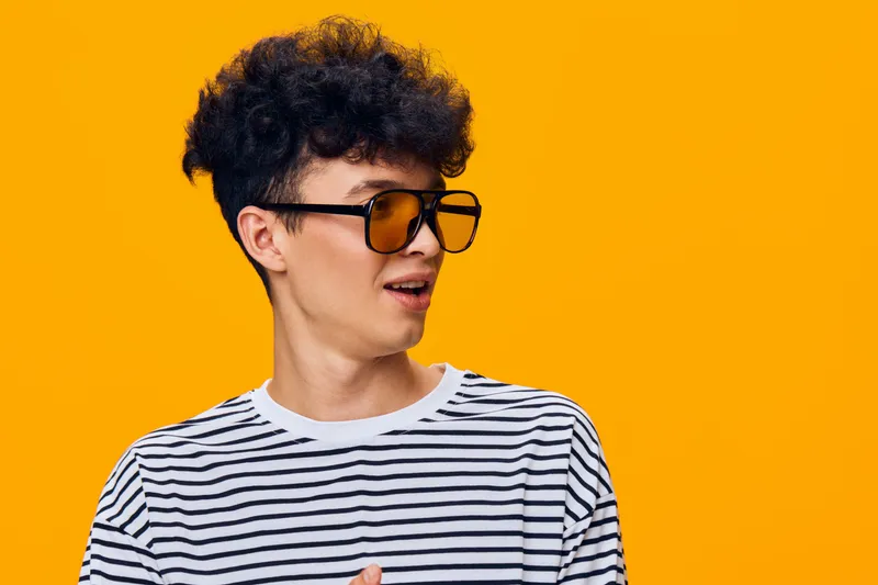 Young man wearing orange sunglasses and a striped shirt looking sideways against a bright orange background, showcasing casual style and confident expression.