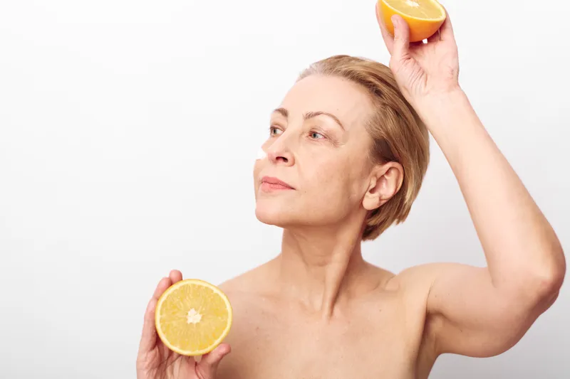 Middle aged woman with short hair holds two halves of a grapefruit near her face on white background. Concept of healthy lifestyle, natural beauty, and skincare with citrus fruit.