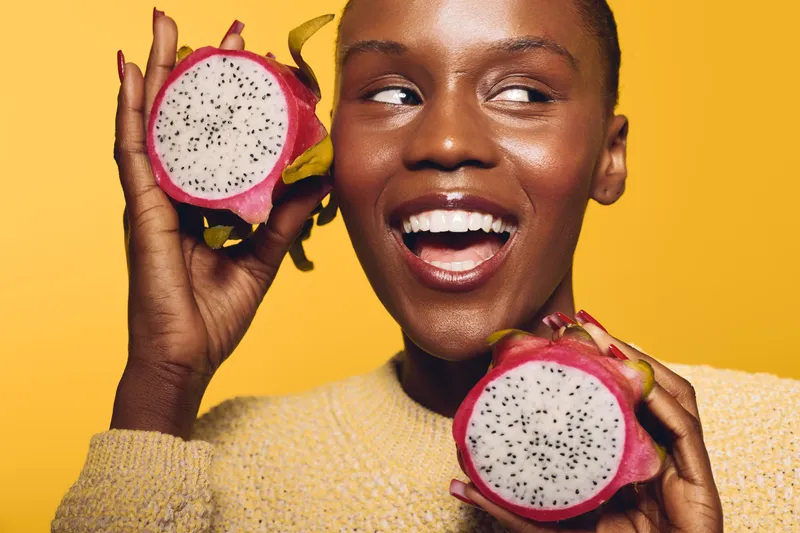 Smiling dark skinned African woman holds two halves of dragon fruit near face on yellow background. Concept for healthy lifestyle, fruit, nutrition, and joyful natural beauty.