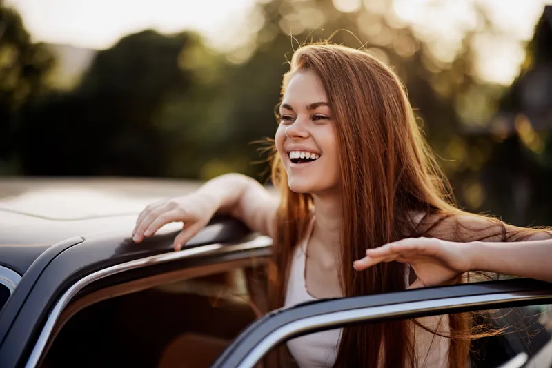 Happy young woman enjoying a road trip, laughing with long hair flowing outside a car window, surrounded by nature in warm sunset light.
