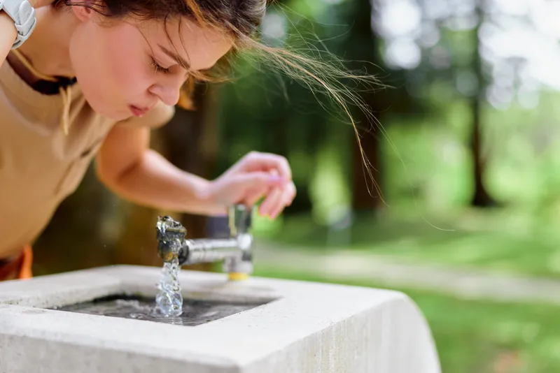 Girl drinking at outdoor fountain, water flowing from tap in park. Child bent over concrete basin, summer scene, casual clothes and focused expression for refreshment in nature.