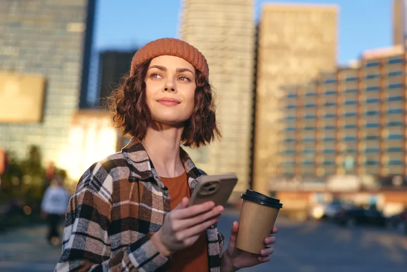 woman coffee smartphone urban beanie candid lifestyle authenticity: Young woman in beanie holds coffee and smartphone on city street at golden hour glow, mindful living portrait with warm sunlight