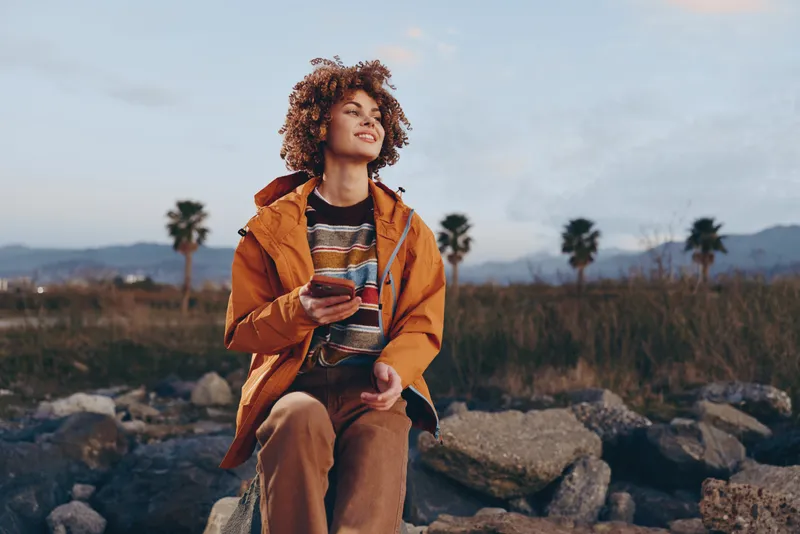 Woman with curly hair in orange jacket outdoors, smiling while walking on rocks, holding a phone, lifestyle travel mood, warm sunset sky, palms in nature