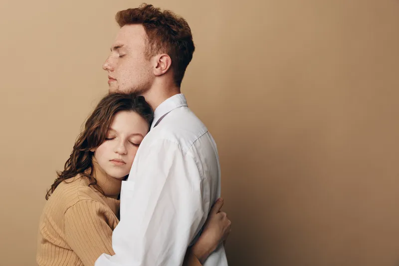 Young couple embraces tenderly with closed eyes against beige background. The woman wears a brown sweater and the man a white shirt. Concept of love, comfort, and emotional support.