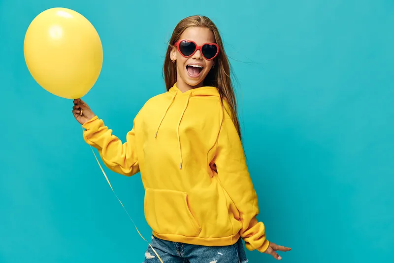 Young woman in yellow hoodie and heart sunglasses smiling with yellow balloon on blue background. Happy girl wearing casual clothes with joyful expression and playful pose.