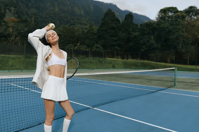 Young female tennis player enjoying a moment on the court, showcasing athletic fashion with a tennis racket and ball, exuding confidence outdoors
