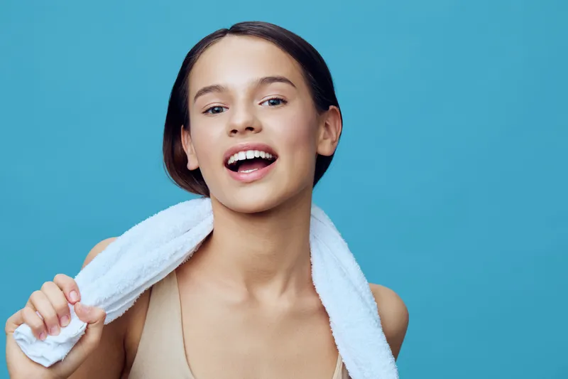 Young female model smiling with a towel on her shoulders against a blue background, conveying freshness, vitality, and a healthy lifestyle