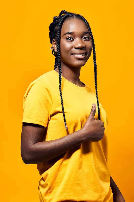 Cheerful young woman with braids wearing a yellow shirt poses against a bright yellow background, expressing confidence and positivity with a thumbs up gesture