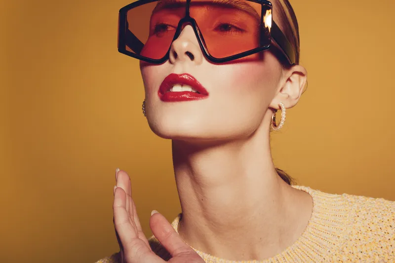 Fashionable woman with red lipstick and oversized orange sunglasses poses against yellow background. She wears hoop earrings and a knitted sweater, showcasing modern style and beauty.