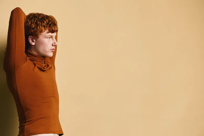 Young man with red hair wearing brown turtleneck stretching arms and looking sideways against beige background with copy space for text.
