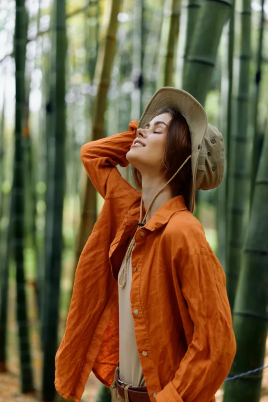 Woman in orange jacket and hat stands among bamboo stalks in a green forest, nature portrait with closed eyes and peaceful expression. Travel mood, outdoor lifestyle scene with soft sunlight.