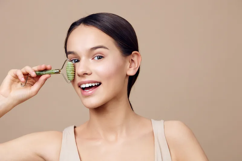 Young woman enjoying facial massage with jade roller, smiling happily against a soft beige background, promoting skincare and self care routines for glowing skin