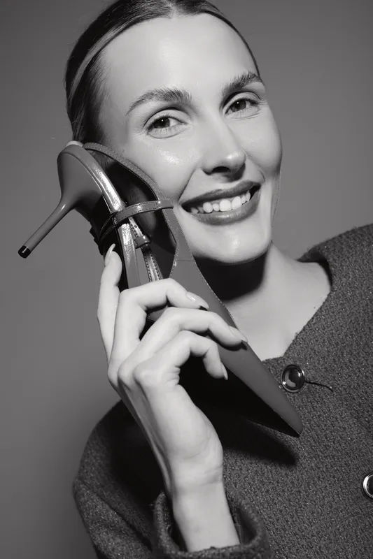 Smiling Caucasian woman with light skin holds high heel shoe near face in black and white portrait. Concept of fashion, happiness, and style expression.