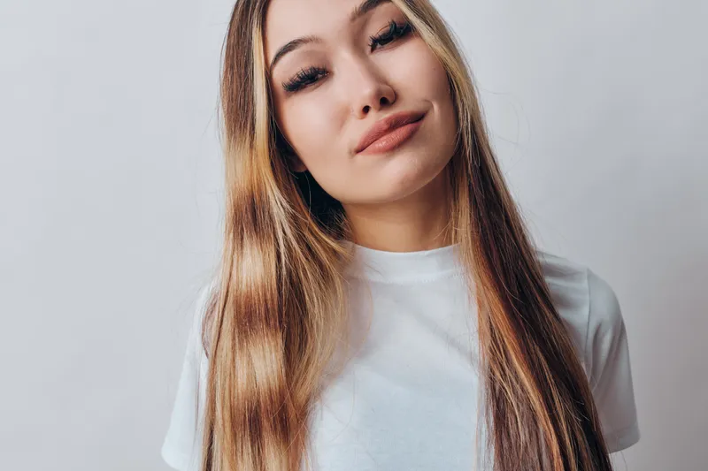 Young woman with long straight hair and natural makeup wearing white t shirt standing against light background. Female model posing with slight smile and relaxed expression.