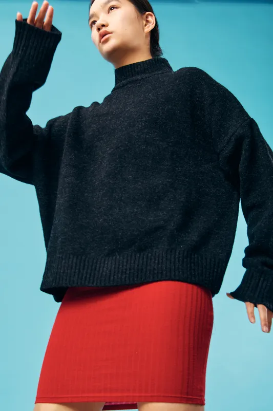 Woman wearing oversized black sweater and red skirt standing against plain blue background with soft lighting in studio. Fashion, casual outfit, and portrait with focus on texture and pose.