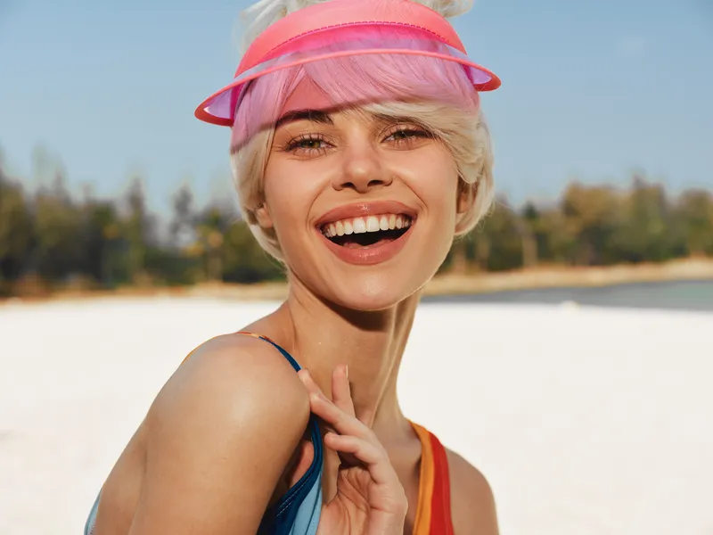 Close up portrait of a joyful woman on a sunny beach wearing a pink visor, bright smile, sunlit skin, playful energy, soft light, vibrant summery mood with blue sky and sea in background.