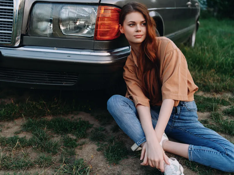 Young woman with long hair in casual outfit sitting next to classic car on grass, evoking a sense of nostalgia and adventure, showcasing timeless style.