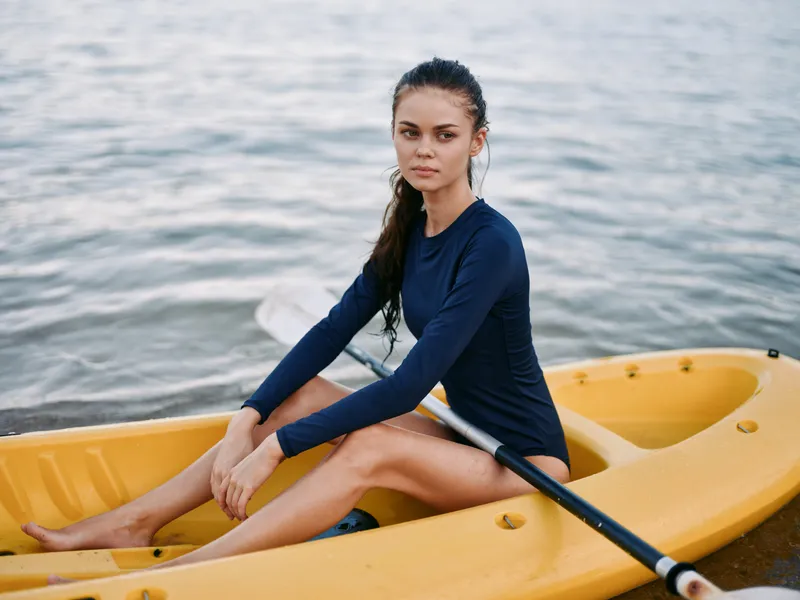 Young woman sits in a yellow kayak on calm water, wearing a navy long sleeve top and bare feet, gazing ahead with a thoughtful, focused expression.