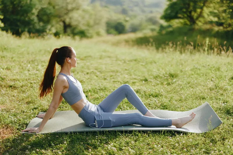Young woman practicing yoga outdoors on a mat in a serene nature setting, wearing comfortable athletic clothing and expressing calmness and relaxation
